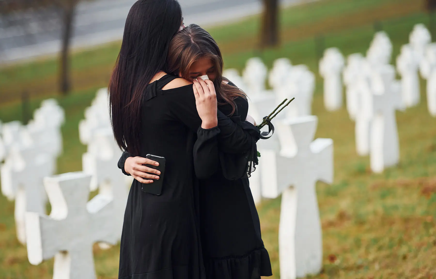 Embracing each other and crying. Two young women in black clothes visiting cemetery with many white crosses. Conception of funeral and death.